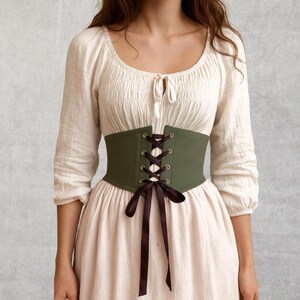 May include: An olive green corset with black lacing and a matching bow. The corset is worn over a cream-colored blouse and a light pink dress. The outfit has a vintage or renaissance style.