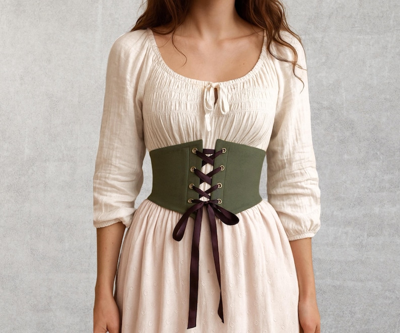May include: An olive green corset with black lacing and a matching bow. The corset is worn over a cream-colored blouse and a light pink dress. The outfit has a vintage or renaissance style.