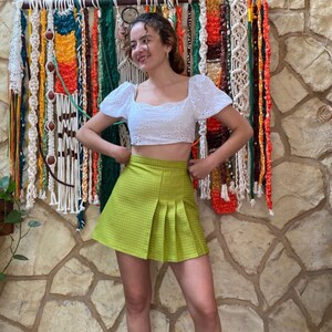 May include: A woman wearing a white eyelet crop top and a lime green pleated skirt. She is standing in front of a wall with a colorful macrame wall hanging.