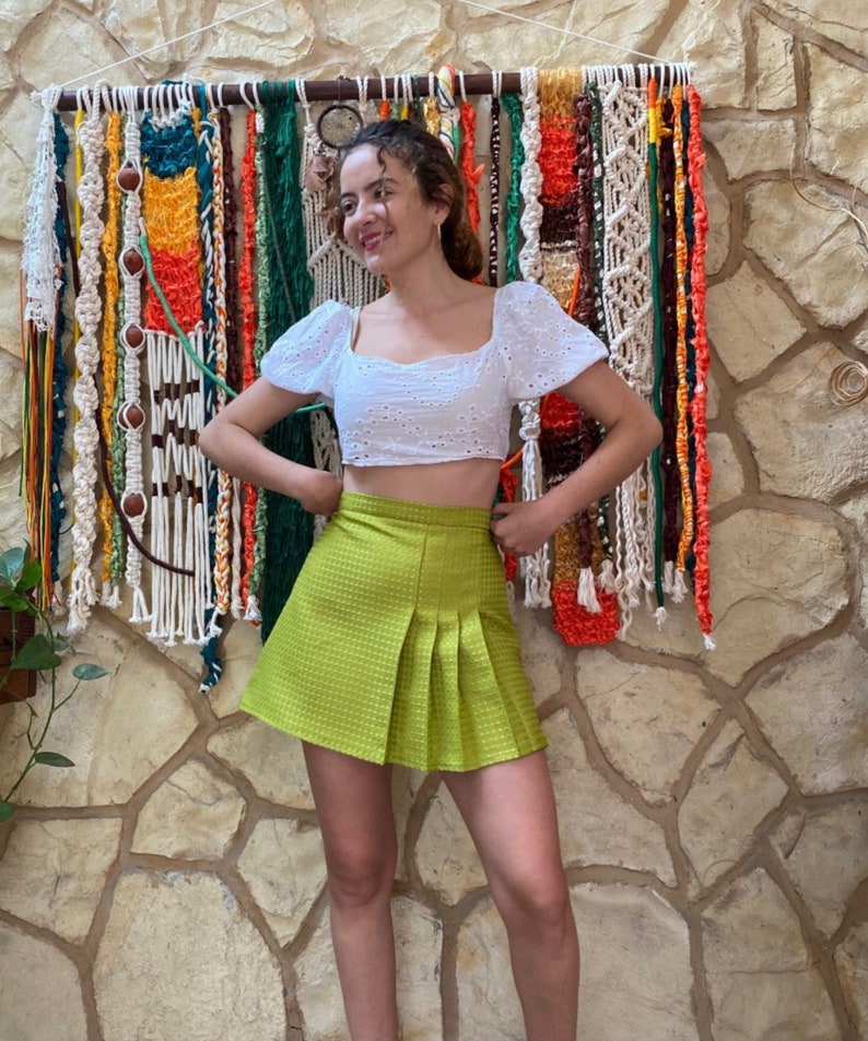 May include: A woman wearing a white eyelet crop top and a lime green pleated skirt. She is standing in front of a wall with a colorful macrame wall hanging.