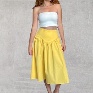 May include: A woman wearing a white tube top and a bright yellow skirt. The skirt has a high waist and a gathered, flared design. The outfit is set against a neutral gray background.