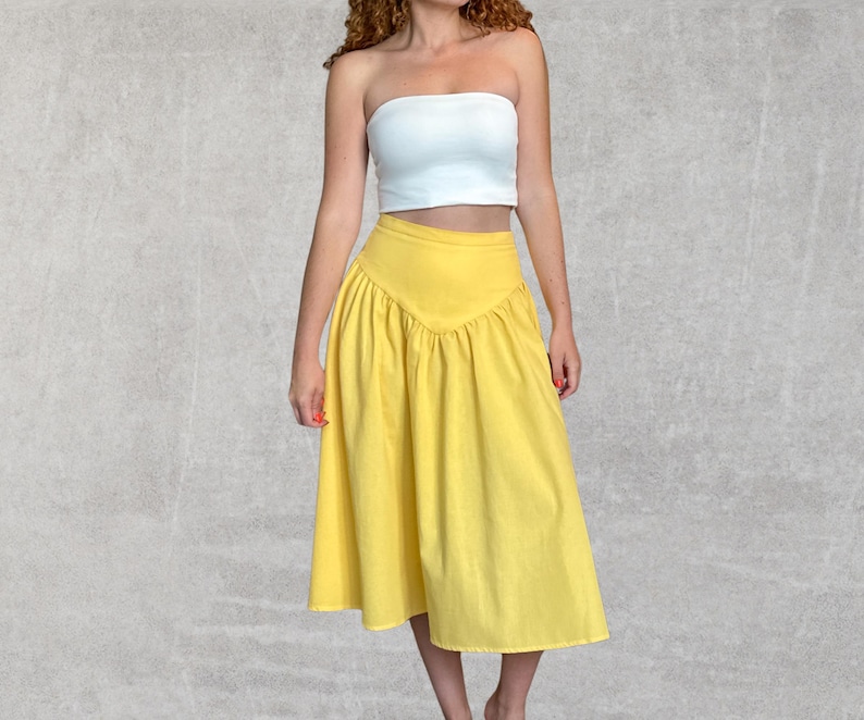 May include: A woman wearing a white tube top and a bright yellow skirt. The skirt has a high waist and a gathered, flared design. The outfit is set against a neutral gray background.