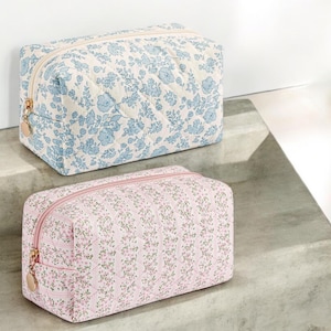 May include: Two quilted fabric cosmetic bags. The top bag is light blue with a white background and a floral pattern. The bottom bag is pink with a white background and a floral pattern.