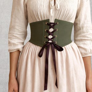 May include: An olive green corset with dark purple ribbon lacing. The corset is worn over a white blouse and a light pink dress. The corset has gold-colored eyelets and a matching dark purple ribbon bow.