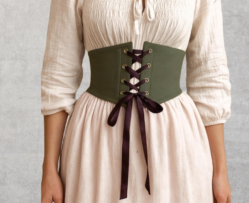 May include: An olive green corset with dark purple ribbon lacing. The corset is worn over a white blouse and a light pink dress. The corset has gold-colored eyelets and a matching dark purple ribbon bow.