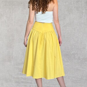 May include: A bright yellow midi skirt with a high waist and a flared silhouette. The skirt is made of a lightweight fabric and has a gathered detail at the waist. The skirt is paired with a white tube top.