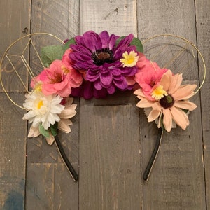 May include: A floral headband with gold wire ears. The headband features a variety of colorful flowers, including pink, purple, and white blooms. The flowers are arranged across the top of the headband, creating a decorative and whimsical accessory.