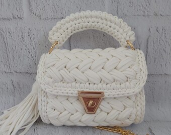 May include: White crocheted handbag with a gold chain strap and a gold clasp. The bag has a tassel and a woven design.