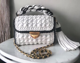 May include: A white crocheted handbag with a black and white braided handle, a gold clasp, and a white tassel. The bag has a gold chain strap.