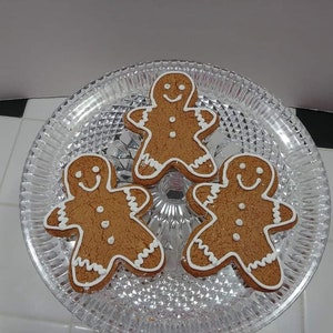 May include: Three gingerbread men cookies on a clear glass cake stand. The cookies are decorated with white icing and have a smiling face.