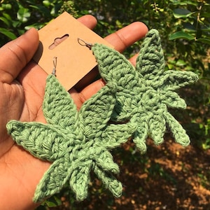 May include: A pair of green crocheted earrings shaped like leaves. The earrings are hanging from silver hooks.
