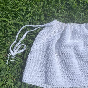 Pode incluir: Uma saia branca de crochê com cintura com cordão. A saia é feita de um tecido texturizado de trama aberta. O cordão é um cordão trançado. A saia é mostrada em um fundo de grama verde.