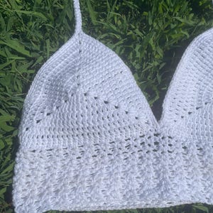 Könnte beinhalten: Ein weißes, handgehäkeltes Oberteil mit Neckholder-Design. Das Oberteil hat ein strukturiertes Stichmuster und einen V-Ausschnitt. Es hat dünne, verstellbare Träger. Das Oberteil ist aus weichem, leichtem Garn gefertigt.