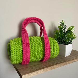 May include: A handmade, lime green crochet bag with bright pink straps. The cylindrical bag is displayed on a wooden shelf, with a small potted plant in the background. The bag's texture and color combination create a vibrant, eye-catching accessory.