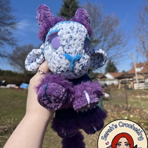 May include: A handmade crochet plushie in shades of purple and white. The plushie has large, round eyes, horns, and fluffy details. The plushie is held up in front of a sunny background. The plushie is a unique, handcrafted item.