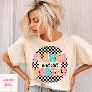 Op de afbeelding: Een wit T-shirt met een zwart-wit geblokt cirkelontwerp. De tekst "Flawed and still worthy" staat in kleurrijke bubbelletters binnen de cirkel.