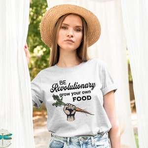 May include: A woman wearing a gray t-shirt with the text "BE Revolutionary grow your own FOOD" and a raised fist holding a carrot. The shirt is a light gray color and the text is in black. The woman is wearing a straw hat and has long brown hair.
