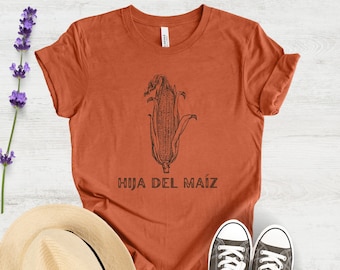 May include: A rust-colored t-shirt with a black and white illustration of an ear of corn and the text "HIJA DEL MAIZ".