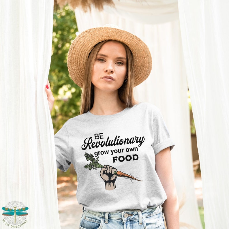 May include: A woman wearing a gray t-shirt with the text "BE Revolutionary grow your own FOOD" and a raised fist holding a carrot. The shirt is a light gray color and the text is in black. The woman is wearing a straw hat and has long brown hair.