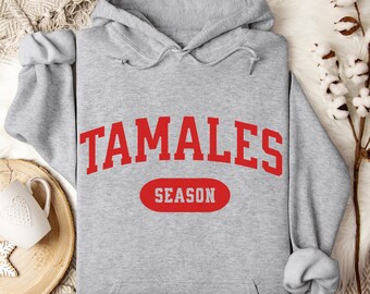 May include: A light gray hoodie with the word "TAMALES" in large red letters across the chest, above a red oval that says "SEASON". The hoodie has a drawstring hood and long sleeves.