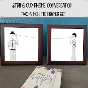 May include: Two framed 6-inch tile prints depicting a man and woman using a string cup phone. The man is wearing a hat and tie, and the woman is wearing a dress. The tiles are framed in dark wood.