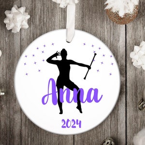 May include: White round Christmas ornament with a purple silhouette of a figure twirling a baton. The ornament has the name "Anna" and the year "2024" printed in purple.