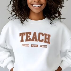 May include: White sweatshirt with the word "TEACH" in brown letters. Below the word are three smaller brown boxes with the words "DIVERSITY", "EQUITY", and "INCLUSION" printed in white.