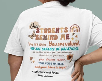 May include: White t-shirt with a brown, orange, and yellow rainbow design and the text "Dear Students Behind Me You are seen. You are valued. YOU ARE CAPABLE OF GREATNESS. No matter where you come from or the color of your skin, your dreams matter, YOUR VOICE MATTERS, and your future is bright! With Love and Pride, Mrs. Johnson"