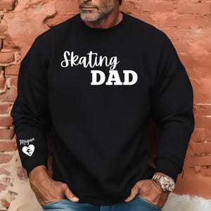 May include: Black long-sleeve sweatshirt with the text "Skating Dad" in white. The left sleeve has a white graphic of a figure skater with the text "Megan" above it.