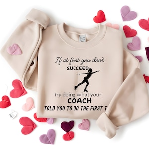 May include: A beige sweatshirt with the text "If at first you don't succeed, try doing what your coach told you to do the first time" and a silhouette of a figure skater. The sweatshirt is surrounded by heart-shaped decorations.