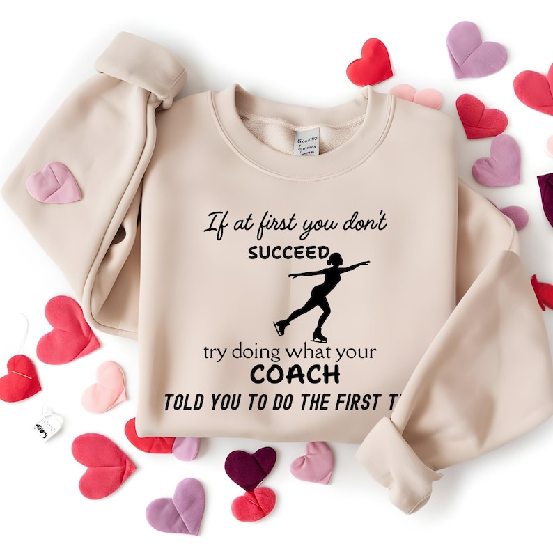 May include: A beige sweatshirt with the text "If at first you don't succeed, try doing what your coach told you to do the first time" and a silhouette of a figure skater. The sweatshirt is surrounded by heart-shaped decorations.
