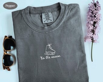 May include: A gray t-shirt with a white graphic of an ice skate and the text "'tis the season". The shirt is folded and placed on a light blue surface, with sunglasses and a purple flower to the side.
