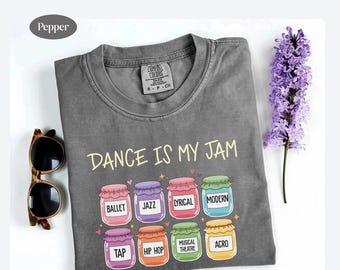 Op de afbeelding: Een grijs t-shirt met de tekst "DANCE IS MY JAM" en kleurrijke potten met dansstijlen zoals Ballet, Jazz en Hip Hop. Een zonnebril en een takje paarse bloemen zijn zichtbaar.