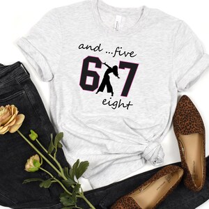 May include: Light grey t-shirt featuring the text "and...five 6 7 eight" in black and pink. The numbers 6 and 7 are in a black box with a dancer silhouette. The shirt is styled with dark denim jeans and leopard print flats.