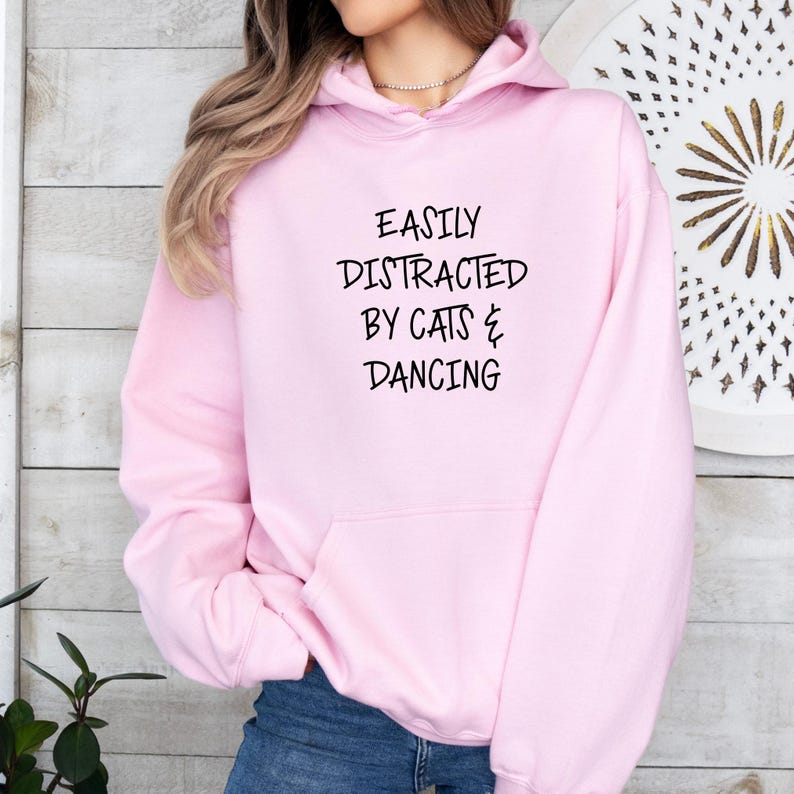 以下が含まれることがあります： 「EASILY DISTRACTED BY CATS & DANCING」の黒い文字が入った、明るいピンク色のパーカー。パーカーには、ドローストリング付きのフードとフロントポケットが付いています。画像はパーカーを着ている人を示しています。