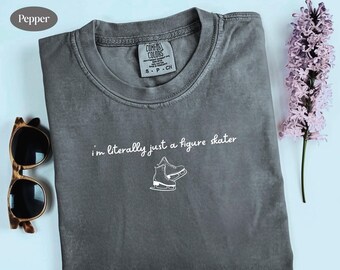 May include: A gray t-shirt with the text "I'm literally just a figure skater" and a graphic of ice skates. The shirt is folded on a light blue surface, with sunglasses and a purple flower stem.