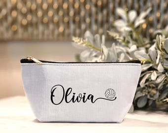 May include: White canvas cosmetic bag with a black zipper and the name "Olivia" in cursive with a volleyball graphic. The bag is a light gray color and has a black trim.