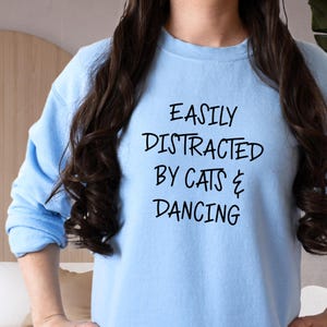以下が含まれることがあります： 「EASILY DISTRACTED BY CATS & DANCING」の黒い文字が入った水色のスウェットシャツ。 画像は上半身のクローズアップです。 スウェットシャツはクルーネックで、柔らかい素材で作られているようです。