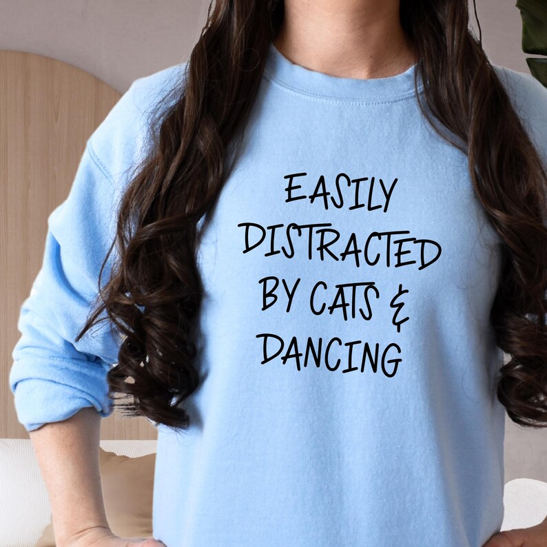 以下が含まれることがあります： 「EASILY DISTRACTED BY CATS & DANCING」の黒い文字が入った水色のスウェットシャツ。 画像は上半身のクローズアップです。 スウェットシャツはクルーネックで、柔らかい素材で作られているようです。