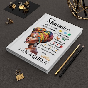 May include: White journal with the name "Shaunice" and the text "I am a Queen." The cover features a portrait of a woman wearing a colorful head wrap. Gold and black pencils and paper clips are on the dark surface.