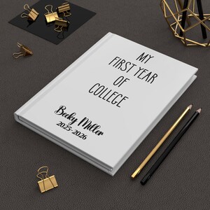 May include: A white hardcover journal with black text that reads MY FIRST YEAR OF COLLEGE, followed by the name Becky Miller and the years 2025-2026. The journal is positioned on a dark surface alongside two pencils and several gold binder clips.