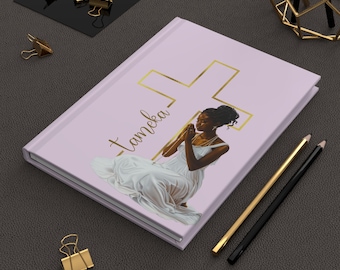 May include: A light purple journal with a gold cross design and the name "Tameka" in gold script. The cover features an illustration of a woman in a white dress praying. Gold and black pencils and gold binder clips are also visible.