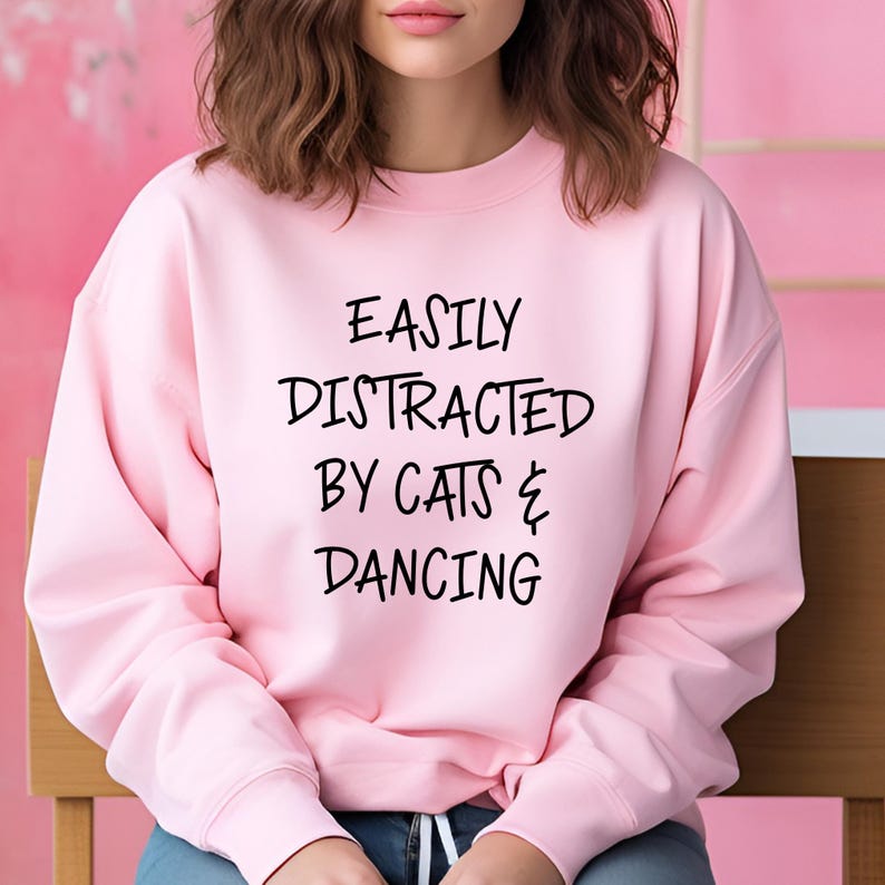 以下が含まれることがあります： 「EASILY DISTRACTED BY CATS & DANCING」の黒い文字が入った、明るいピンク色のスウェットシャツ。クルーネックのスウェットシャツは柔らかい生地で作られています。背景は明るいピンク色で、木製の椅子が見えます。