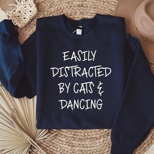 以下が含まれることがあります： 「EASILY DISTRACTED BY CATS & DANCING」の白文字がプリントされたネイビーブルーのスウェットシャツ。クルーネックのスウェットシャツは、帽子や装飾品を背景に、織り込まれた表面に表示されています。