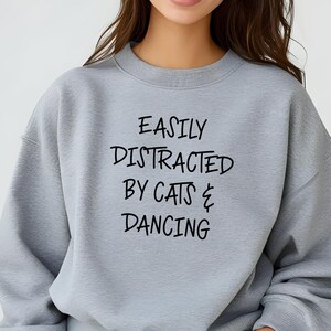 以下が含まれることがあります： 「EASILY DISTRACTED BY CATS & DANCING」の黒い文字がプリントされた、ライトグレーのスウェットシャツ。クルーネックと長袖のデザインです。生地は柔らかく快適に見えます。