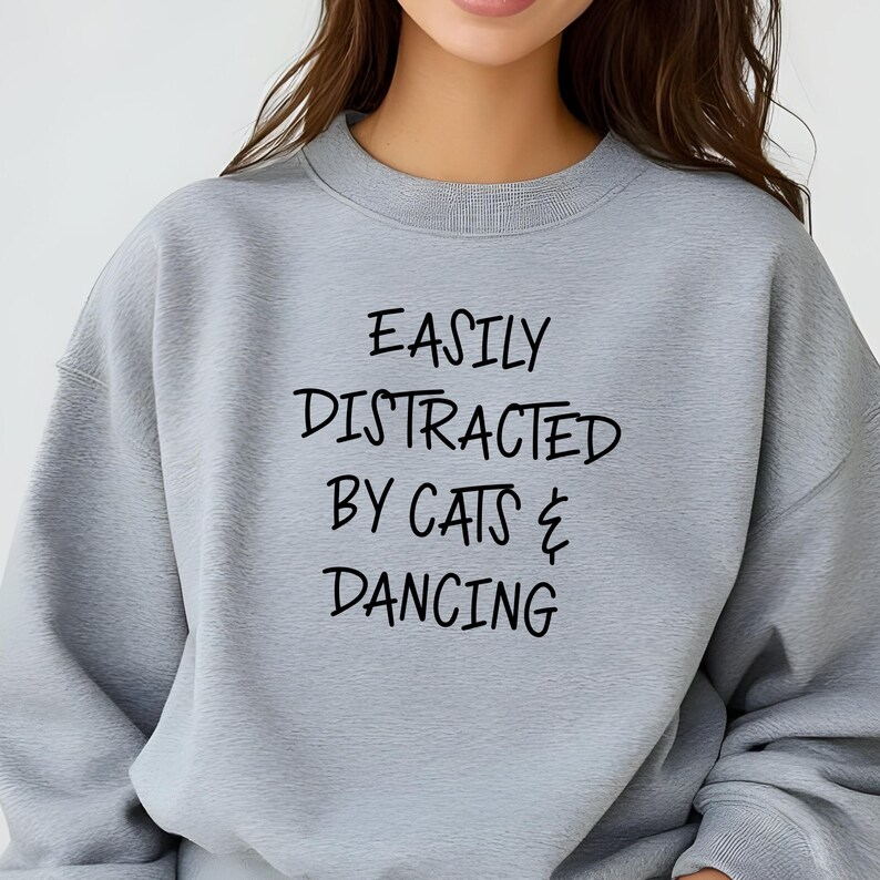 以下が含まれることがあります： 「EASILY DISTRACTED BY CATS & DANCING」の黒い文字がプリントされた、ライトグレーのスウェットシャツ。クルーネックと長袖のデザインです。生地は柔らかく快適に見えます。
