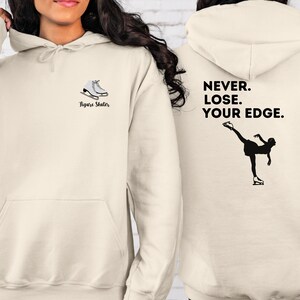 May include: A cream-colored hoodie with a figure skater silhouette and the text "Never. Lose. Your Edge." on the back. The front of the hoodie has a pair of ice skates and the text "Figure Skater".
