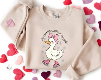 May include: Beige sweatshirt with a graphic of a cartoon goose wearing pink ice skates and a pink bow. The text "Silly Skater Goose on the Loose" is printed above the goose. Scattered around the sweatshirt are heart-shaped decorations in various colors.
