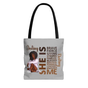 Pu&ograve; includere: Una borsa tote grigia con un manico nero. La borsa presenta un'illustrazione marrone di una donna con il testo "She is" in grandi lettere marroni. Il testo "Destiny" appare due volte sulla borsa, una volta in marrone e una volta in oro. Le parole "Brave, Fierce, Giving, Strong, Fearless, Blessed, Beautiful, Powerful, Confident, Enough, Me" sono scritte verticalmente in lettere dorate.