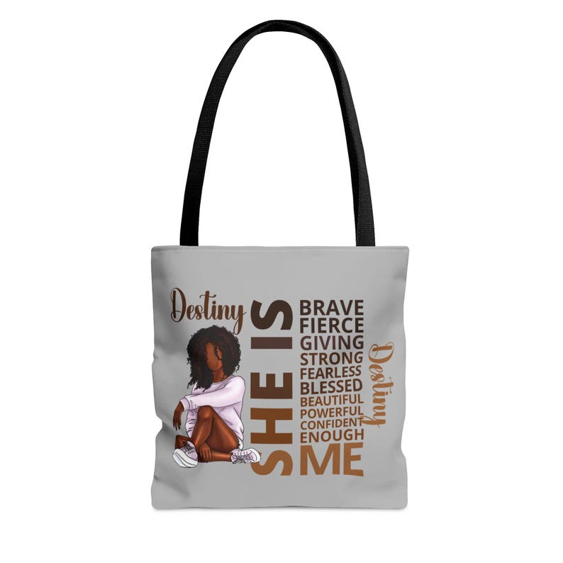 Pu&ograve; includere: Una borsa tote grigia con un manico nero. La borsa presenta un'illustrazione marrone di una donna con il testo "She is" in grandi lettere marroni. Il testo "Destiny" appare due volte sulla borsa, una volta in marrone e una volta in oro. Le parole "Brave, Fierce, Giving, Strong, Fearless, Blessed, Beautiful, Powerful, Confident, Enough, Me" sono scritte verticalmente in lettere dorate.